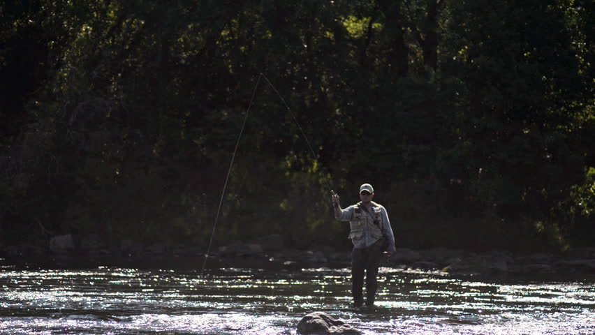 A wide shot of a man in waterproof overalls as he stands in a river and casts his lure with his rod while he spends his day fly fishing.