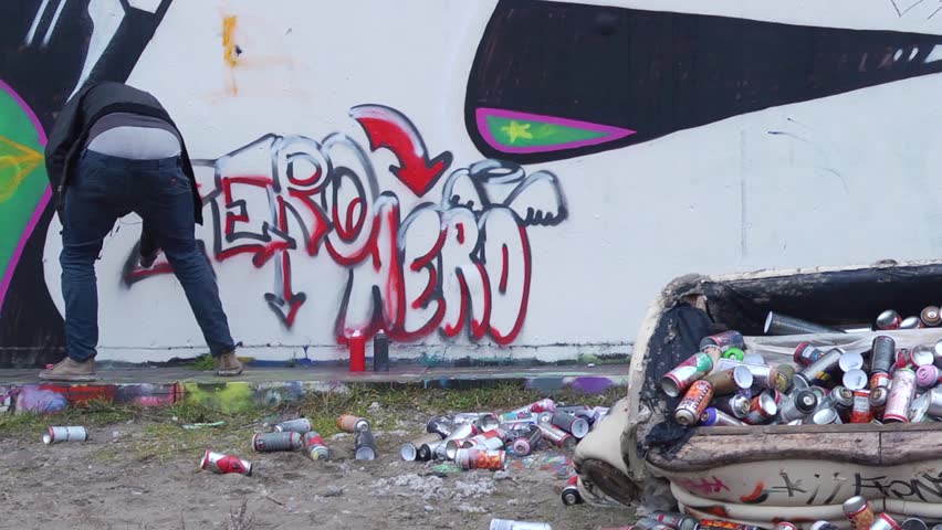Pan over a graffiti wall, towards an artist at work, who is interrupted by law enforcement, and runs away, bailing out