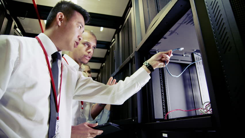 Three people of mixed ethnicity are working in a data center with rows of server racks and super computers. They are walking up and down and checking all of the equipment. 