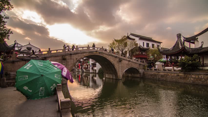 SHANGHAI - CIRCA 2013: Time Lapse Cloudy Sunset of an old Chinese water town with traditional bridge old architecture. People traffic on a bridge by the River in Qibaozhen, China