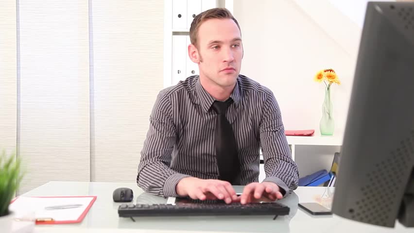 Young businessman seated at his desk concentrating on typing on a desktop computer keyboard, then picking up phone and talking
