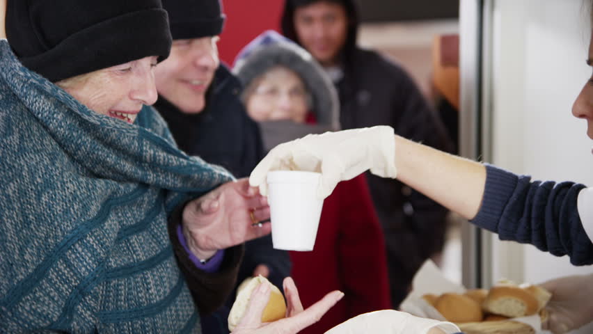 Friendly voluntary workers standing at a soup kitchen serving hatch are handing out cups of hot soup and bread to a waiting line of homeless and needy people.