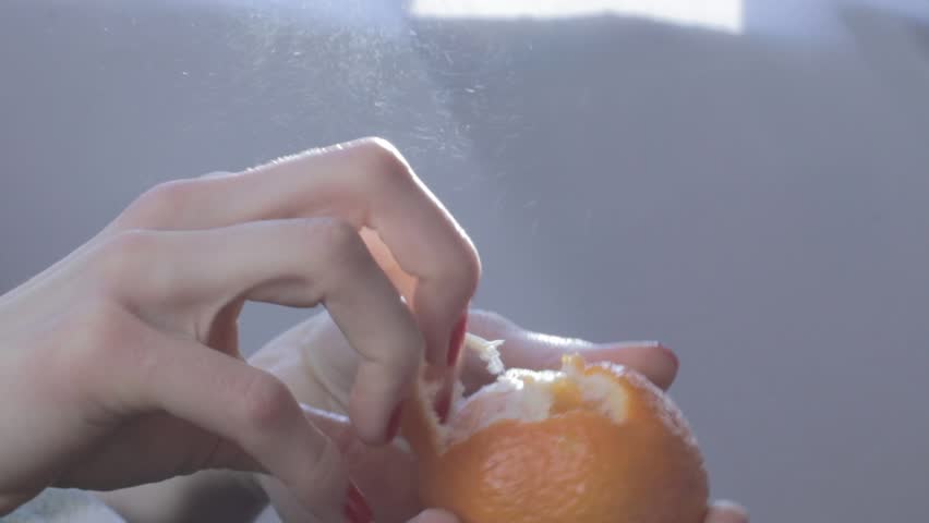Hands of young woman peel tangerine (mandarin). Orange fruit splashes are fontaining to air and well seen through sun rays. Background is neutral grey. Feminine hands have nice manicure with red nails