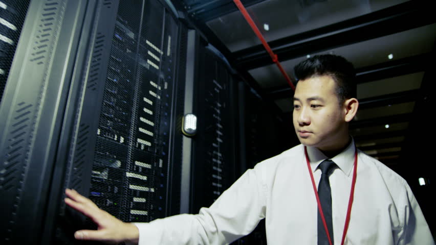 A young male IT engineer of Asian ethnicity is working in a data center with rows of server racks and super computers. He is looking into data cabinets and checking cables and other equipment. 