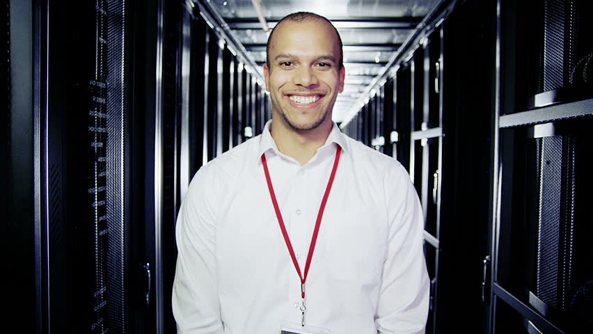 Portrait of a male IT engineer who is working in a data center with rows of server racks and computers. 