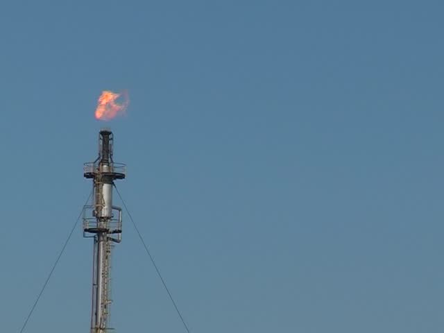 Gas flares and burn off from a chimney at a fuel plant on a sunny day against a blue sky, 