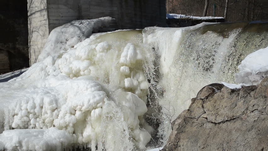old river water dam and water flow mumur between frozen ice icicles in winter.
