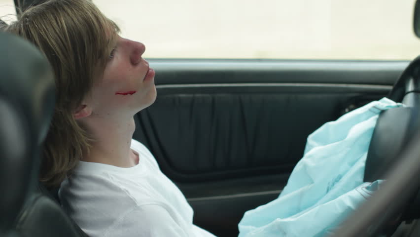 Teenage driver regains consciousness and can be seen through the side window of his wrecked and smoking car. Recorded with camera on jib arm.