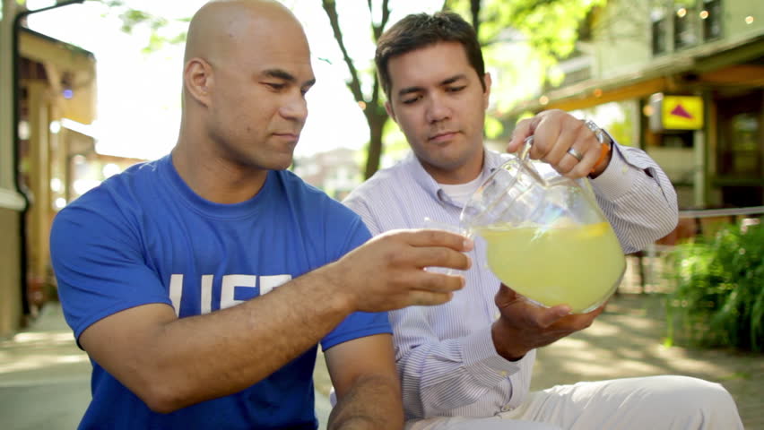 Man in a blue T-shirt that says "LIFE" sits on the street with a bowl of lemons. He hands one to another man who sits next to him and who pours them both lemonade.