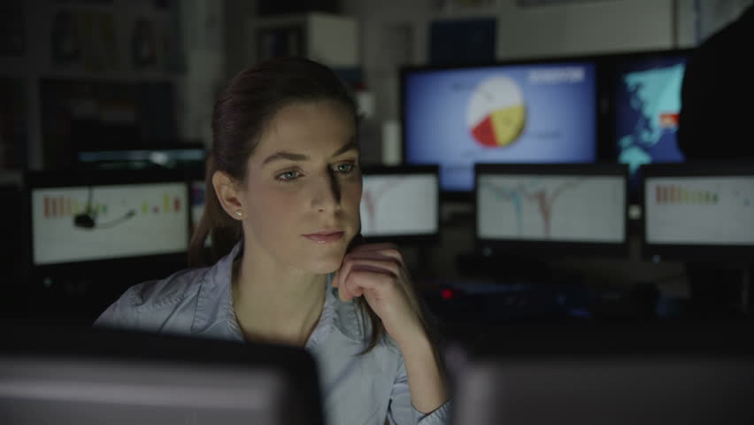 A young business woman is working late at night in a dark office filled with computer screens. She is feeling tired and overworked.