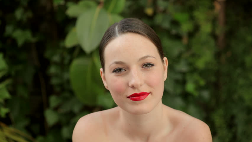 Close up portrait of a young beautiful woman with bare shoulders, wearing bright red glossy lipstick and smiling outdoors, in a green forest background.