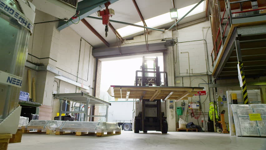 A worker in a warehouse is using a forklift truck to move empty wooden pallets around. Sunlight streams into the space through the open shutter door.