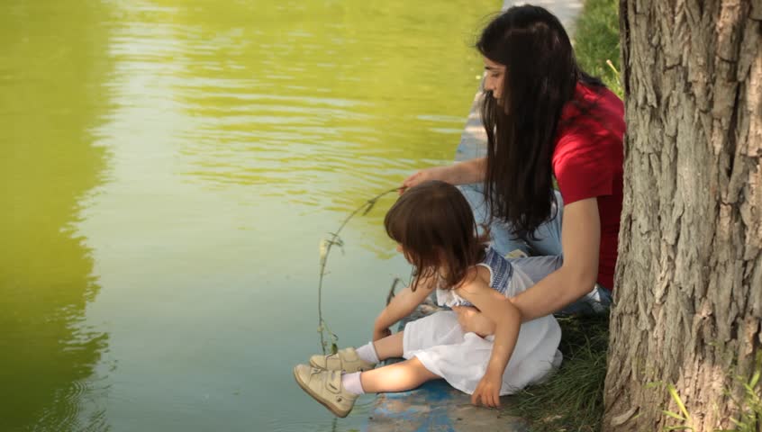 Brunette young adult mother teaches little baby daughter fishing.
