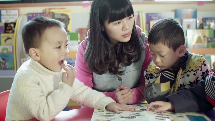 asian kindergarten teacher and her students sit at a small table reading a book together in a classroom in china. dolly shot. 