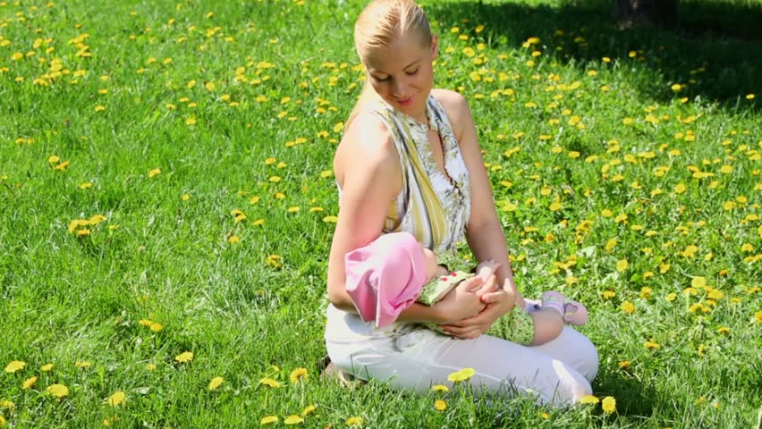 Young woman holds baby on hands while sits at lawn of dandelions
