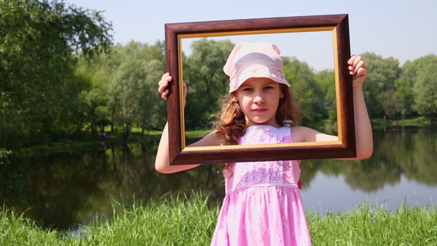 Little girl raises picture frame and his head passes through it, near pond at summer park