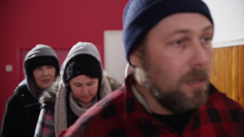 A group of homeless and needy people, wrapped up warm against the cold outside are standing in line in a refuge or soup kitchen. In slow motion.