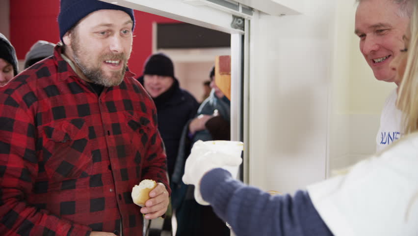 Friendly voluntary workers standing at a soup kitchen serving hatch are handing out cups of hot soup and bread to a waiting line of homeless and needy people. 