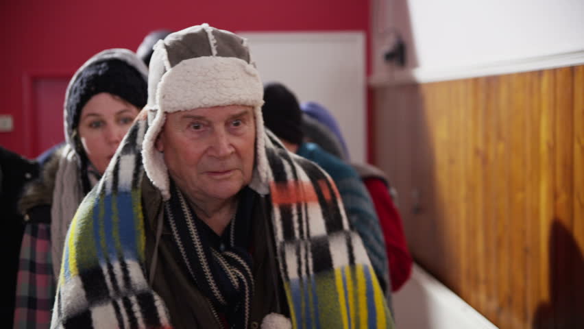 A group of homeless and needy people, wrapped up warm against the cold outside are standing in line in a refuge or soup kitchen. In slow motion.