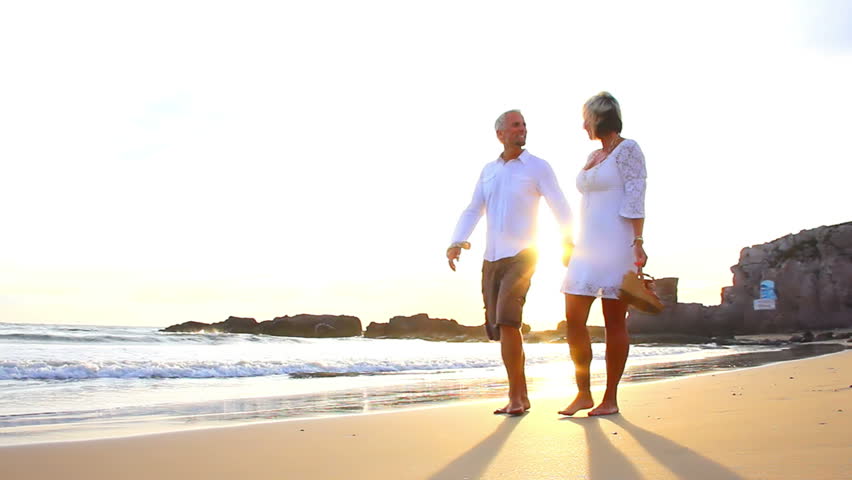 An older couple holds hands and walks down the beach at sunset getting their feet wet