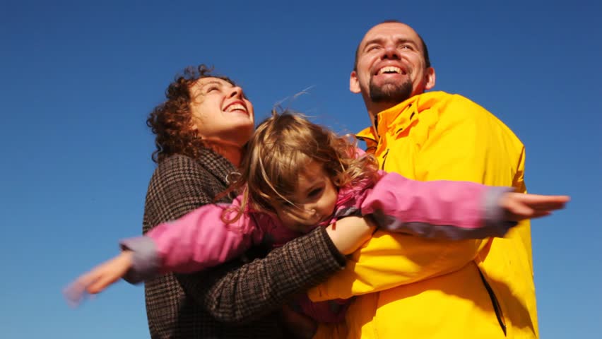 mum and daddy shakes daughter on hands and all of them smile against clear sky 