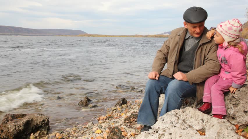 daddy with daughter sit on stone on riverbank, look afar and talk 