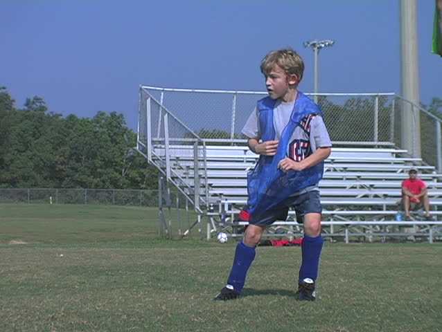 Young boy chases a soccer ball, kicks it then trips with another player.  Both fall.