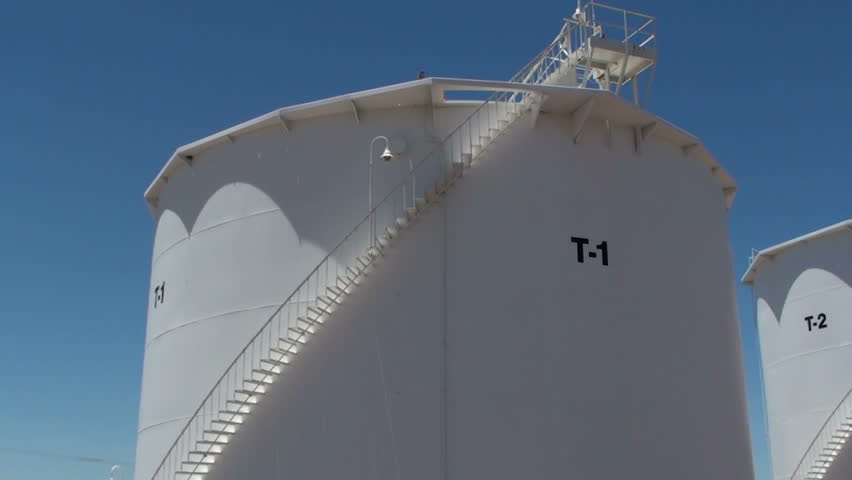 Pan right, row of oil storage tanks, stark white against blue sky.