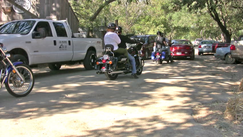Video of motorcycles leaving Luckenbach Texas. Large group of motorcycle riders with Harley. Large oak trees and small country store and environment. 