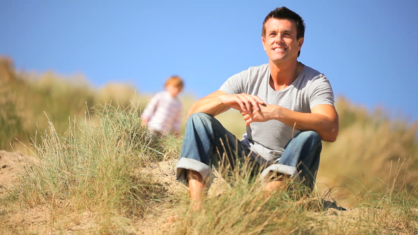 Adorable little blond boy & his young father enjoying time together  sitting in sand dunes by the coast