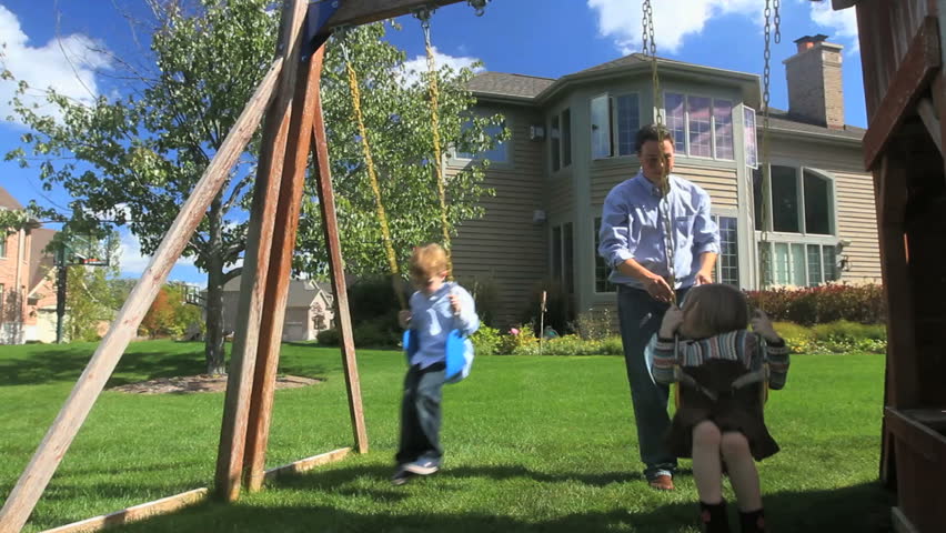 A father pushes his two kids on the swings in the back yard of their luxury home 