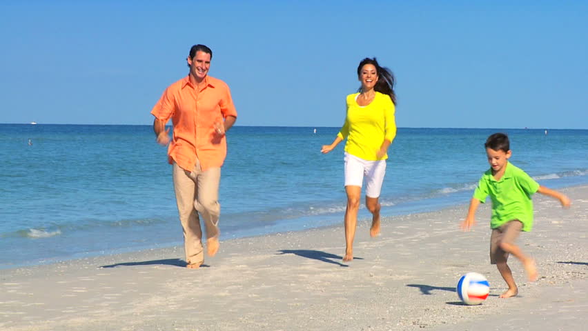 Young family having fun together kicking a ball along the beach filmed 60FPS