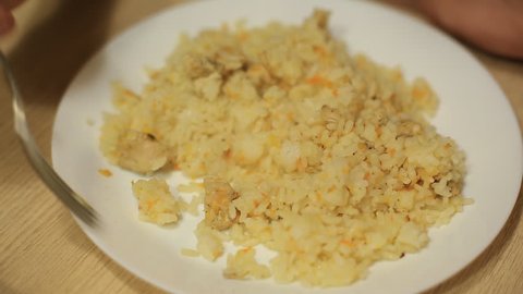 a man eating rice with a fork with chicken meat from a white plate at home at a wooden table.