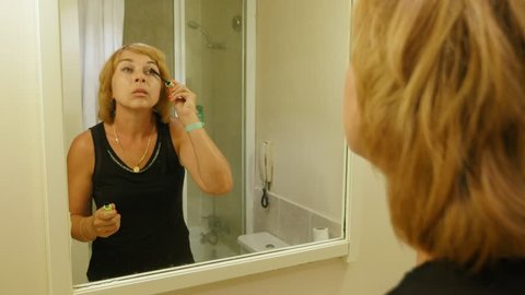 Portrait Of A Beautiful Blond Woman With Bob Haircut Imposing Makeup On Her Long Eyelashes With A Special Thin Black Brush Before A Mirror Indoors
