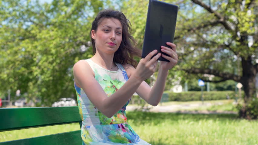 Pretty girl sitting in the park and doing selfies on tablet , HD stock footage clip